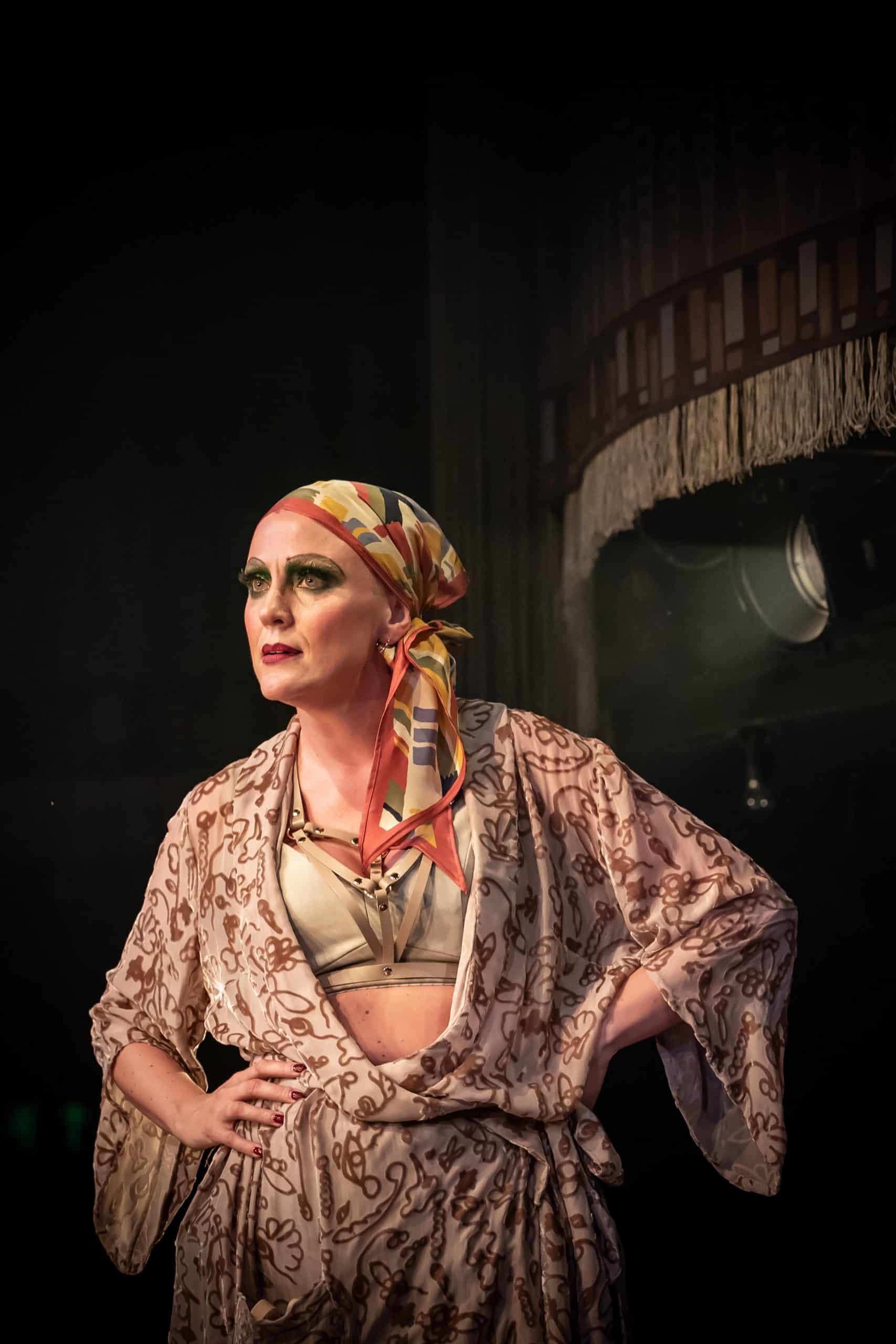 Jessica Kirton as Fraulien Kost in Cabaret at the Kit Kat Club, London. She wears a colourful headscarf and patterned dressing gown