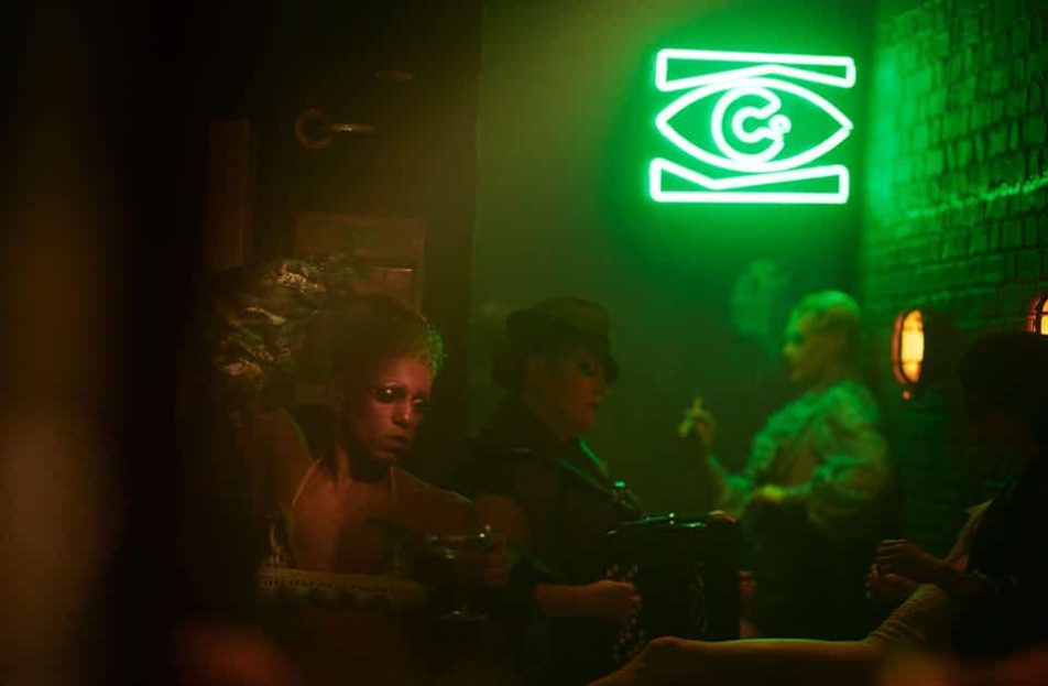 Inside the Kit Kat Club Broadway. The KKC eye as a green neon sign is on the back wall. A group of zany characters stand around the bar drinking and smoking